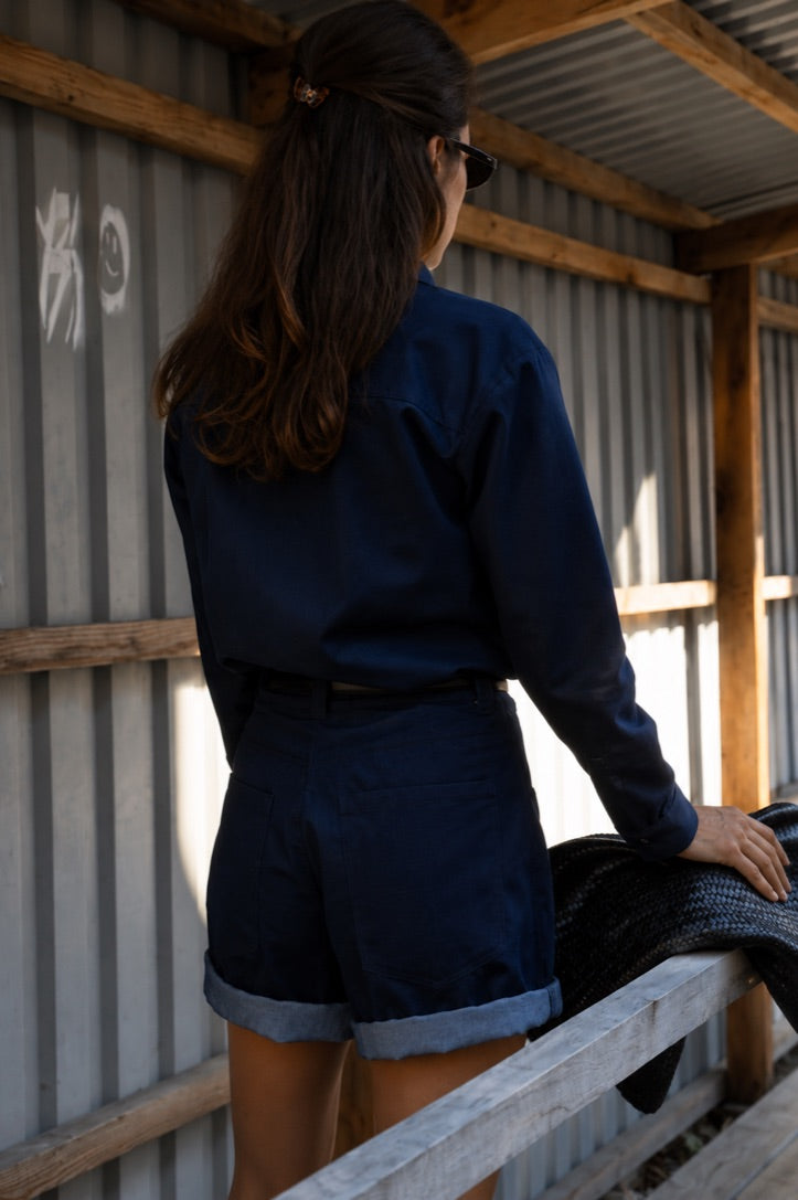 A woman viewed from behind wearing a dark navy blue long-sleeve button-up shirt and matching navy shorts with cuffed hems, standing in front of a corrugated metal wall.