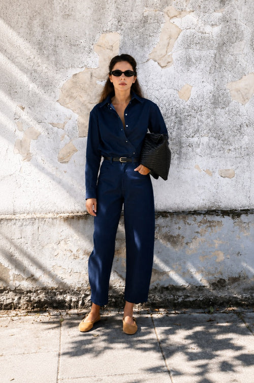 A woman stands outdoors against a weathered concrete wall wearing a navy blue long-sleeve button-up jumpsuit with a thin black belt, tan loafers, black sunglasses, and holding a black textured shoulder bag.