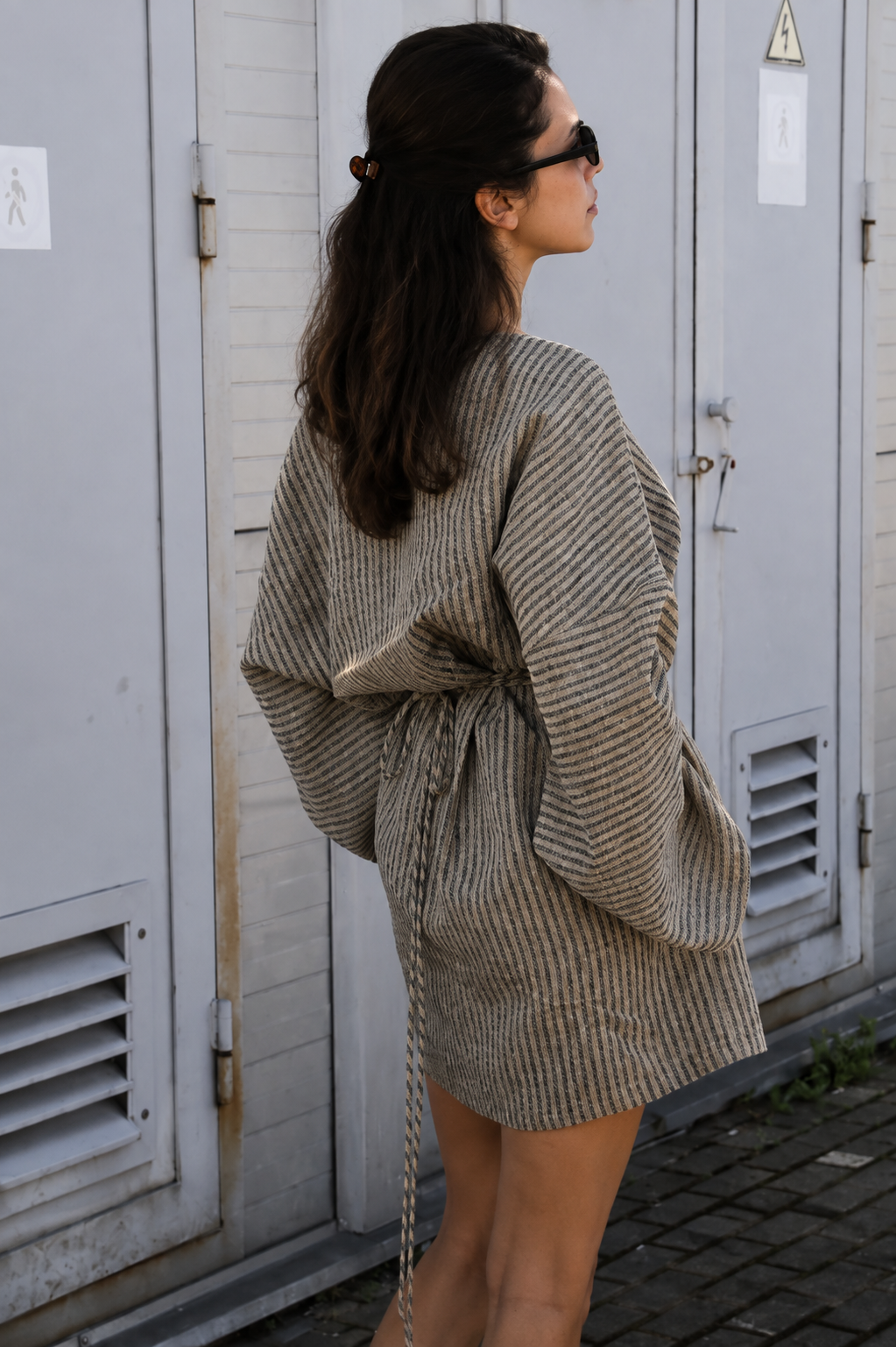 A woman wearing a beige and brown striped wrap jacket with a tie waist, photographed from the back against a background of gray utility sheds.