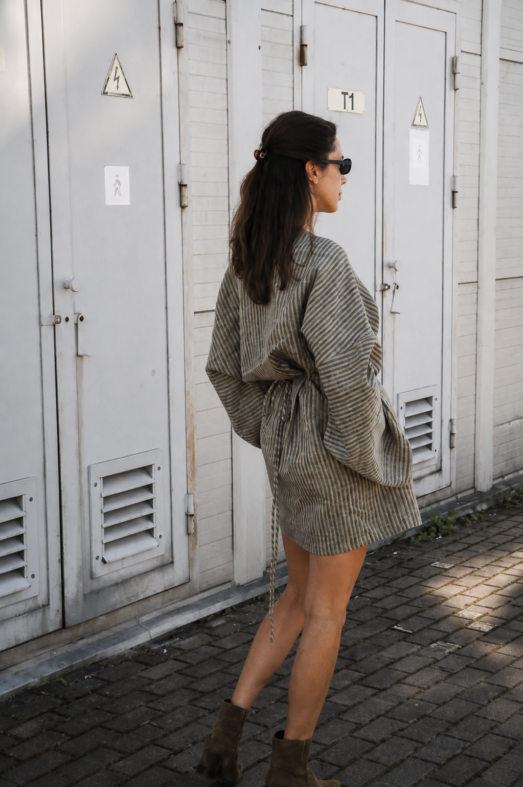 A woman wearing a brown and beige vertically striped wrap-style mini dress and brown suede ankle boots walks past a row of white storage lockers outdoors.