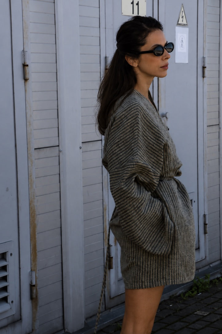 A woman wearing a textured gray and beige herringbone knit belted wrap dress or robe, styled with black sunglasses, standing in front of white wooden shutters.