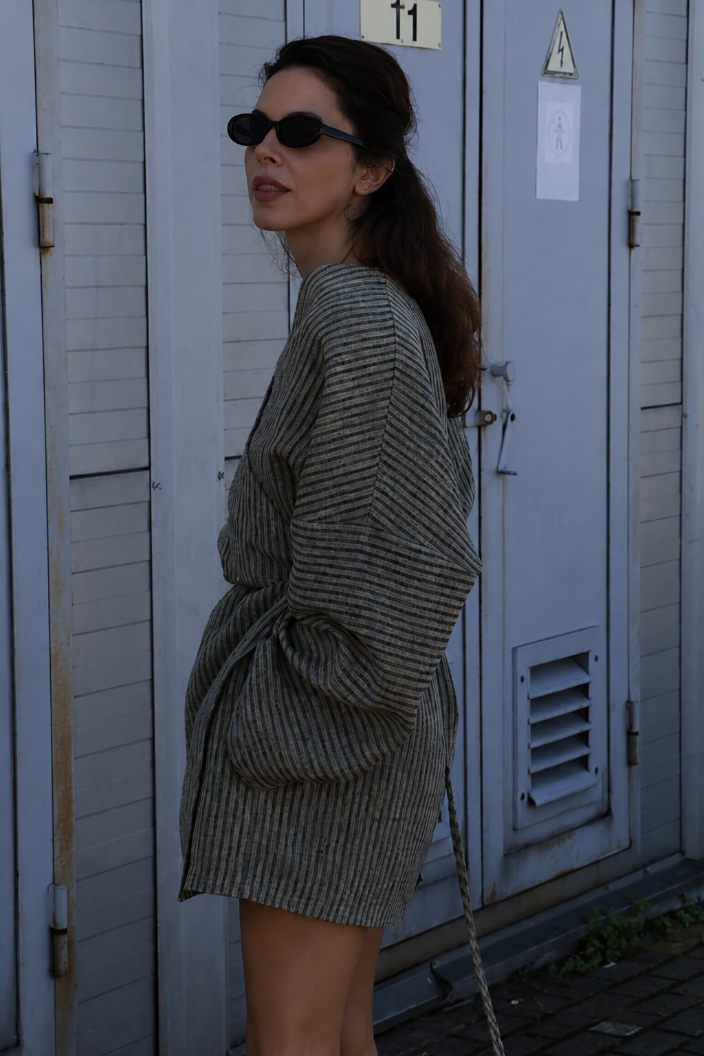 A woman wearing a gray and black vertically striped oversized mini dress with a tie waist, styled with black sunglasses, standing in front of gray metal utility doors.