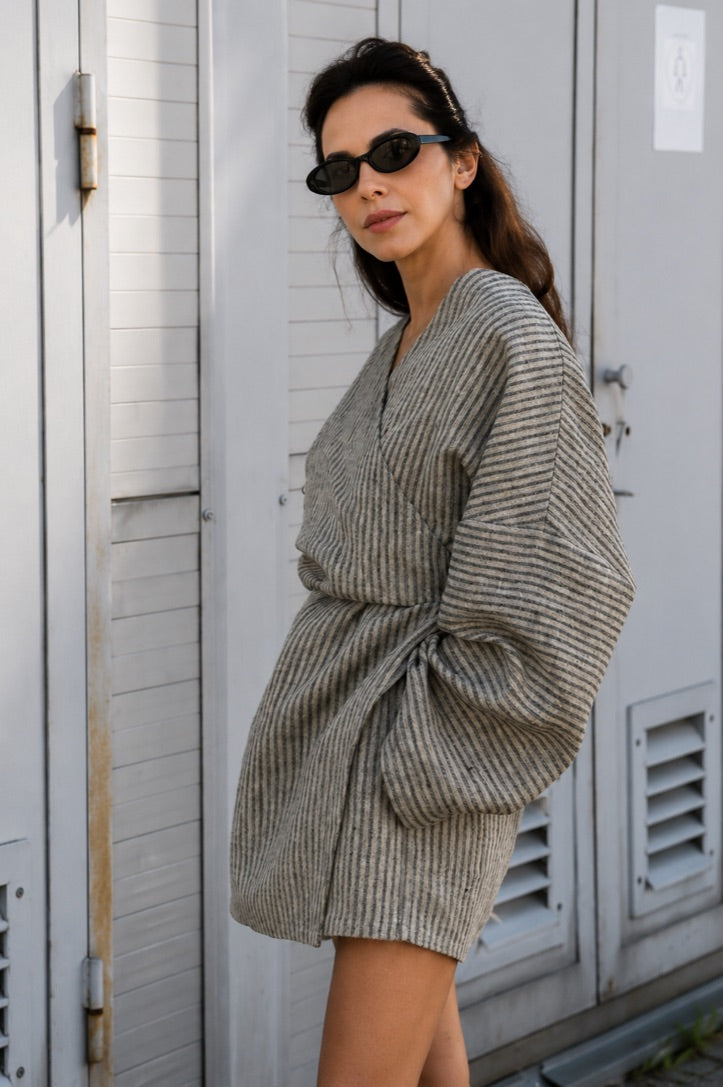 A woman wearing a gray and white vertically striped wrap-style mini dress with oversized sleeves, standing outdoors in front of white shutters.
