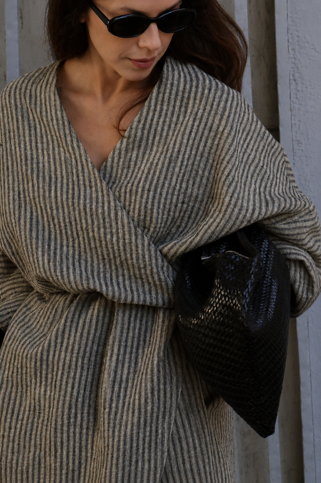 A woman wearing a thick, striped beige and gray wrap shawl or poncho, styled with black sunglasses and a black woven handbag, set against a neutral gray wooden background.