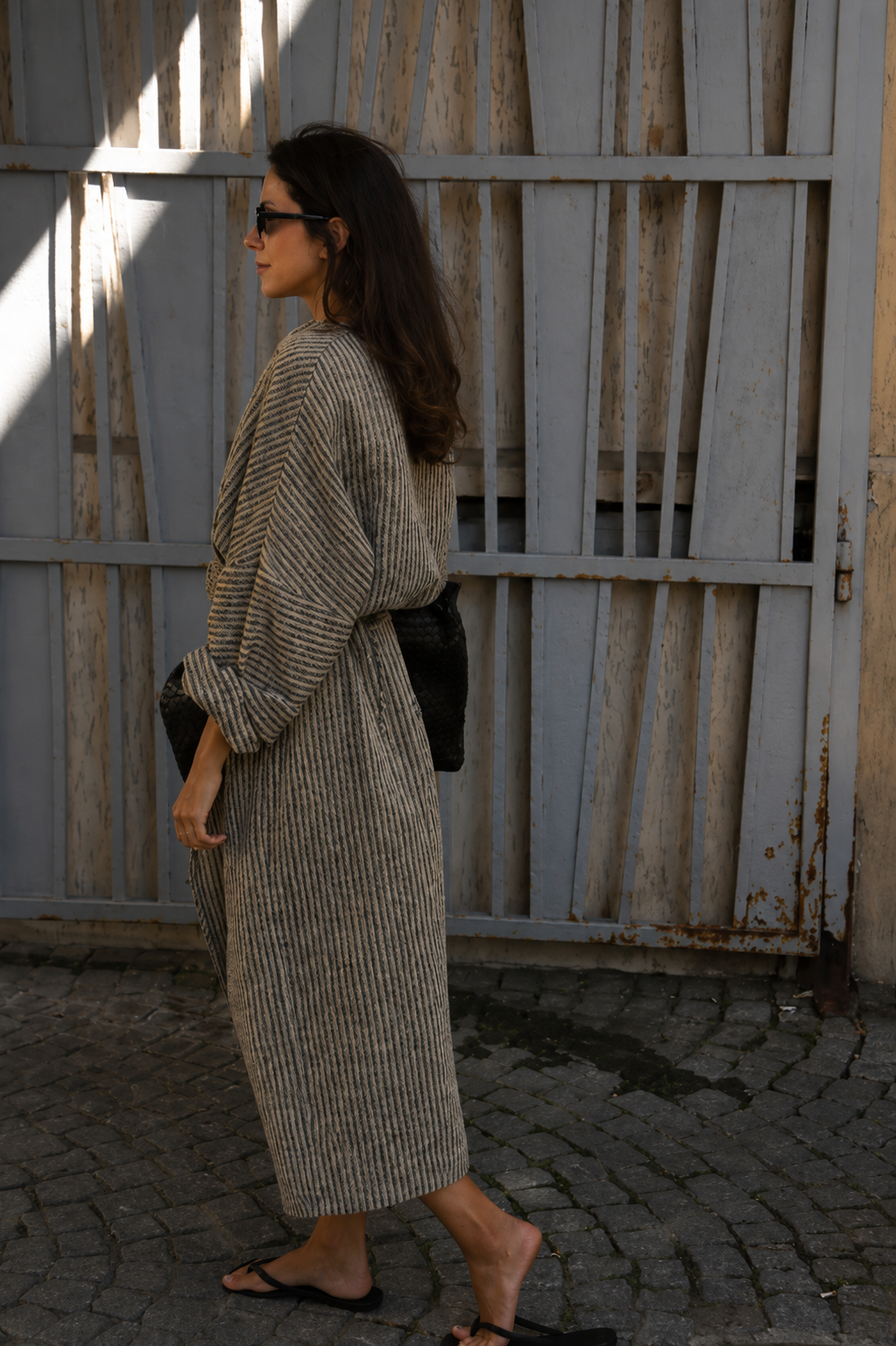 A woman wearing a long beige and gray vertically striped knit robe walks past a weathered wooden gate outdoors, carrying a black shoulder bag.