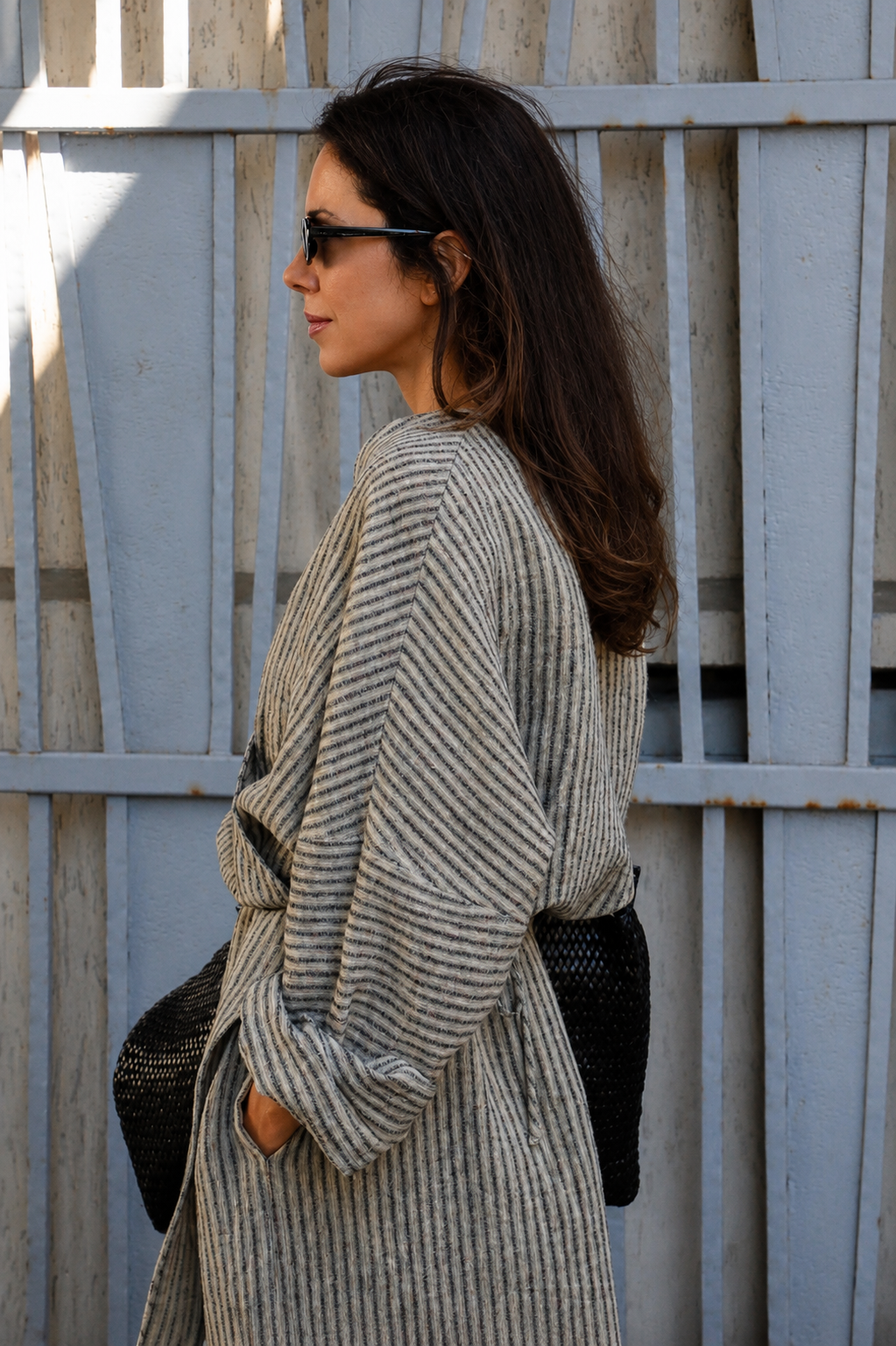 A woman in side profile wearing a beige and gray vertically striped belted coat, black sunglasses, and carrying a black woven shoulder bag, standing in front of a weathered light blue wooden fence.