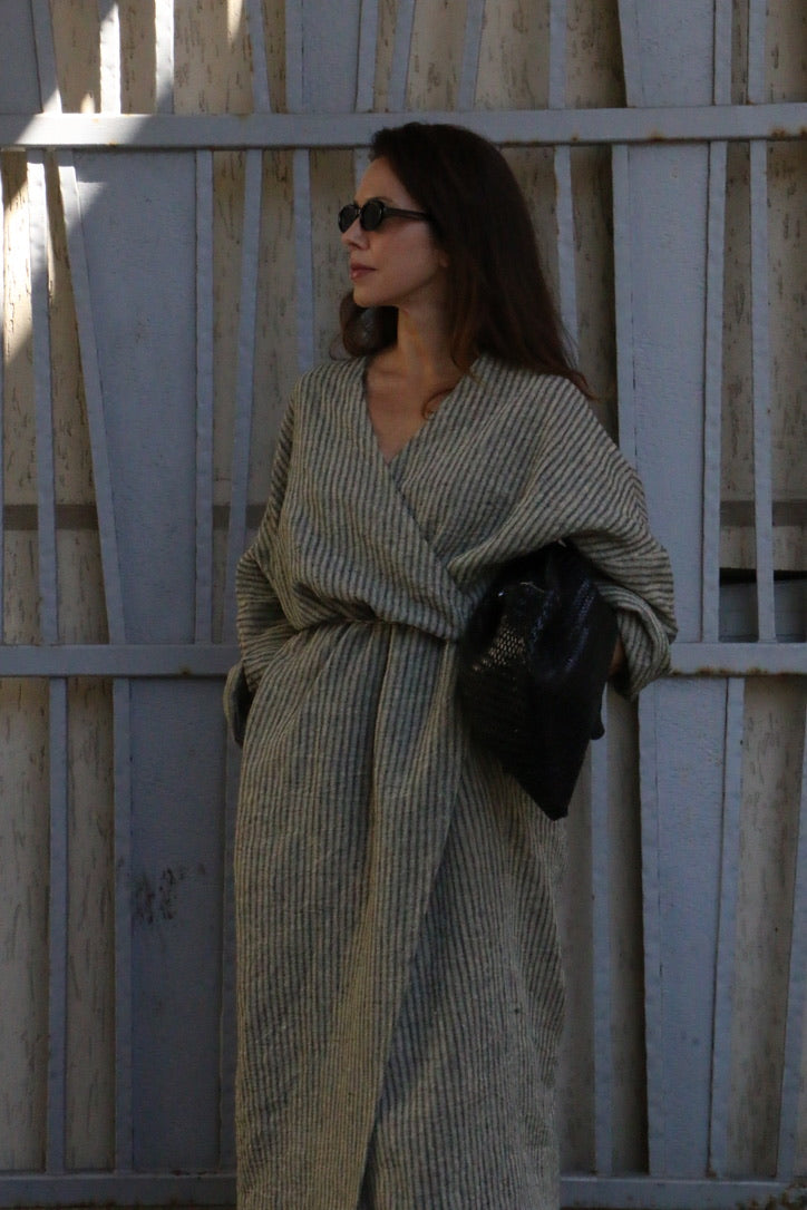 A woman wearing a long, loose, striped beige and gray wrap coat or robe, holding a black clutch bag, standing in front of a weathered gray wooden gate.