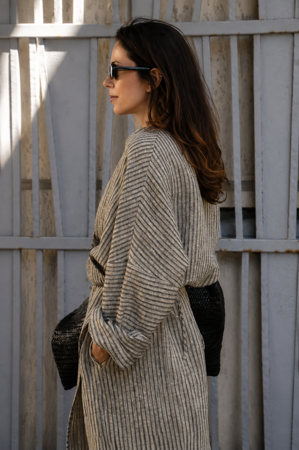 A woman in side profile wearing a beige and gray vertically striped knit cardigan, black sunglasses, and a black woven handbag, standing in front of a weathered gray wooden fence.