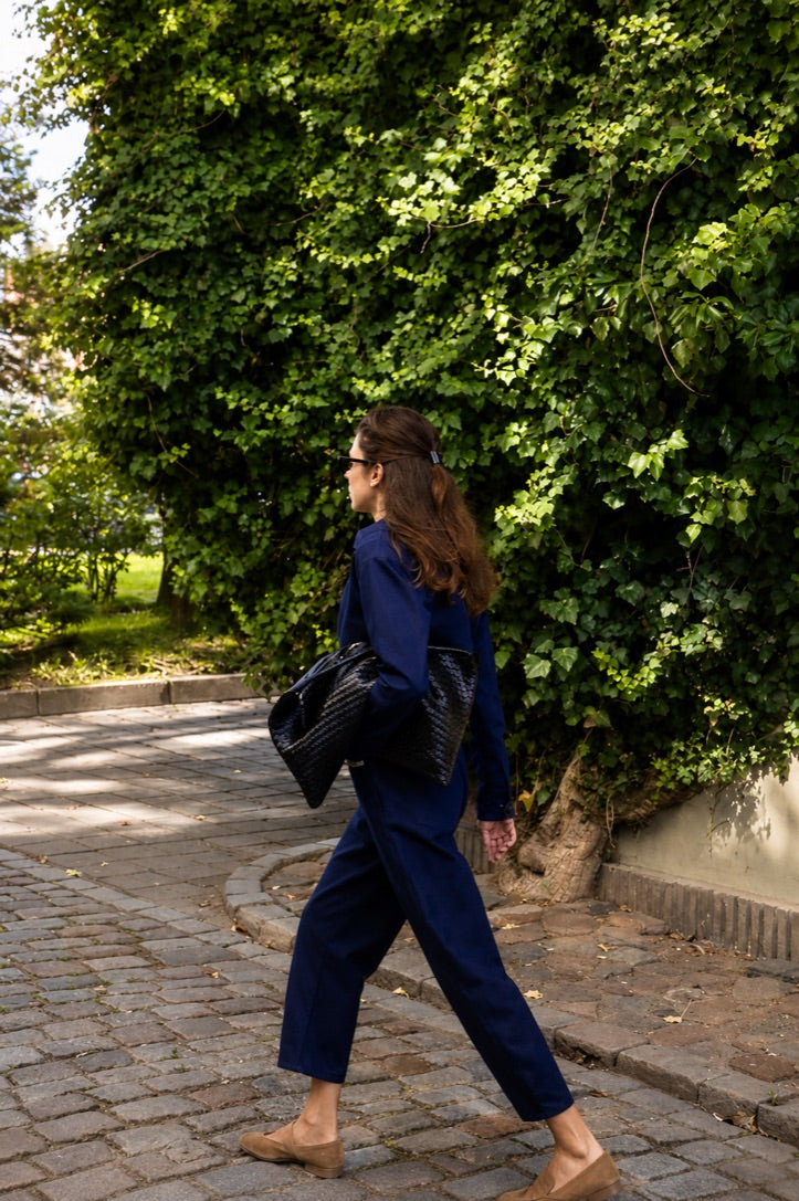 A woman walking outdoors on a cobblestone path wearing a navy blue jumpsuit, carrying a black textured shoulder bag.