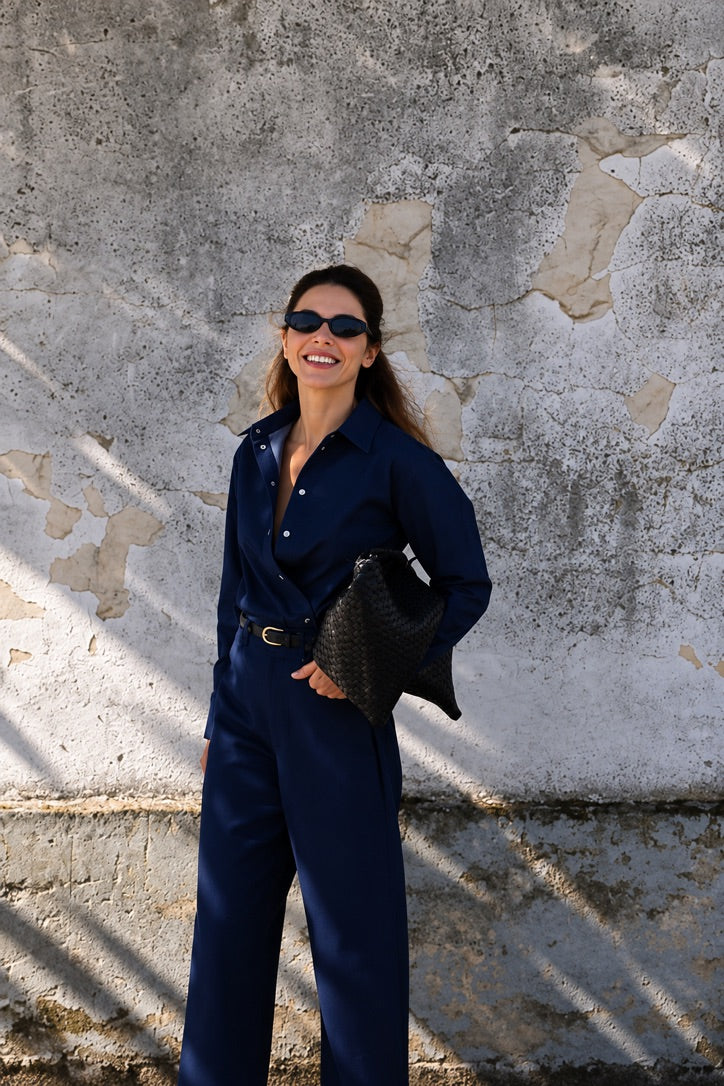 A woman wearing a navy blue button-up shirt and matching wide-leg trousers, styled with a black woven clutch and sunglasses, posing against a textured concrete wall.