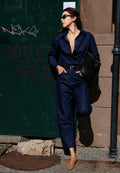 A woman wearing a dark navy blue satin jumpsuit, black sunglasses, and tan suede shoes stands on a city street next to a graffiti-covered green door, holding a black textured handbag.