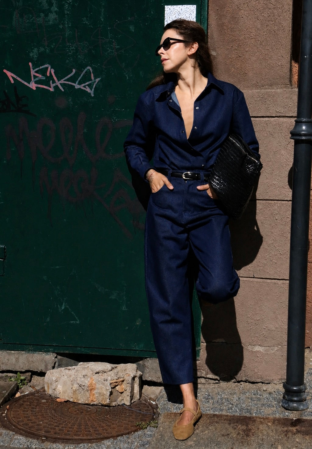 A woman wearing a dark navy blue satin jumpsuit, black sunglasses, and tan suede shoes stands on a city street next to a graffiti-covered green door, holding a black textured handbag.